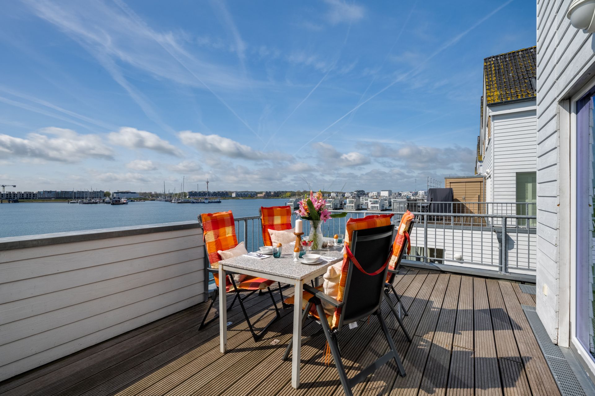 Terrasse mit gedecktem Tisch und Stühlen, Blick auf das Wasser und Gebäude im Hintergrund.