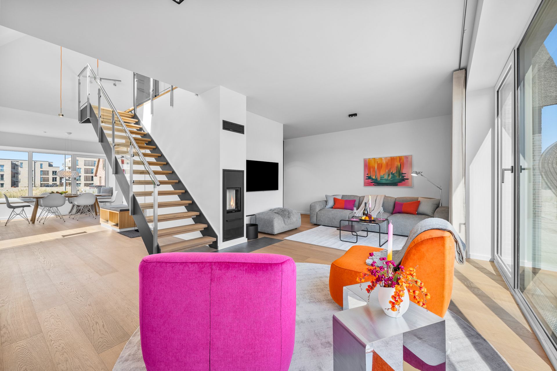 Modernes Wohnzimmer mit bunten Sesseln, Sofa, Fernseher und Treppe, große Fenster lassen viel Licht herein.
