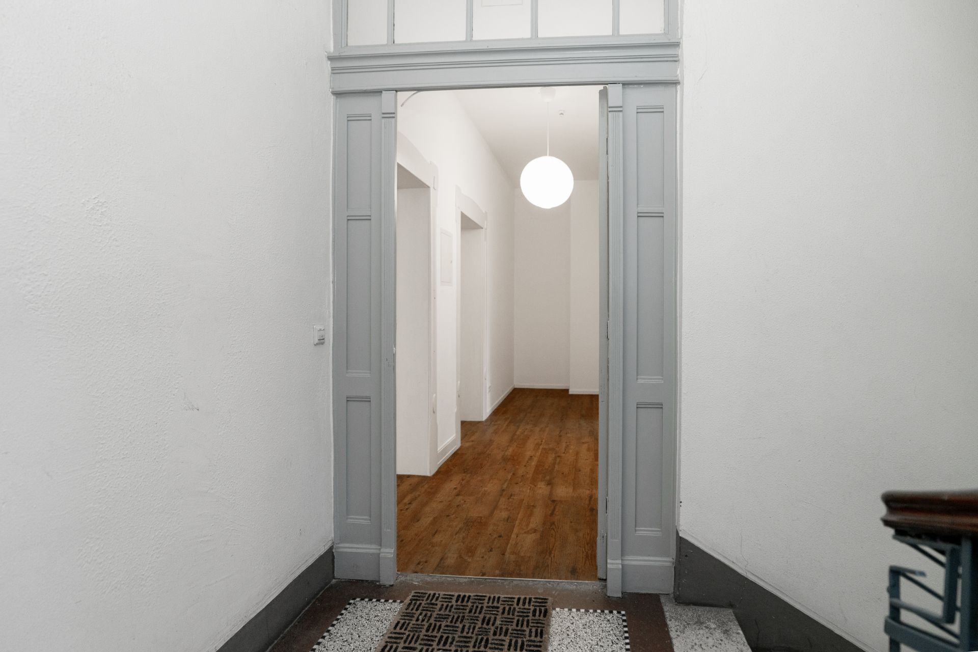 Eingang zu einem Flur mit Holzfußboden und weißer Wand, runde Lampe an der Decke.