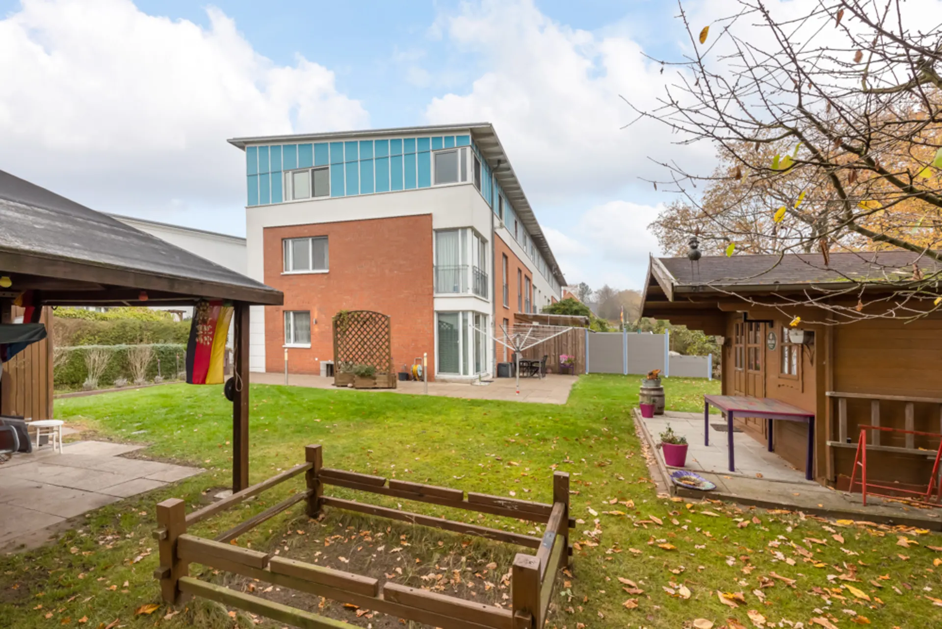 Garten mit Holzhaus, Rasen, kleiner Terrasse und modernem Wohngebäude im Hintergrund.