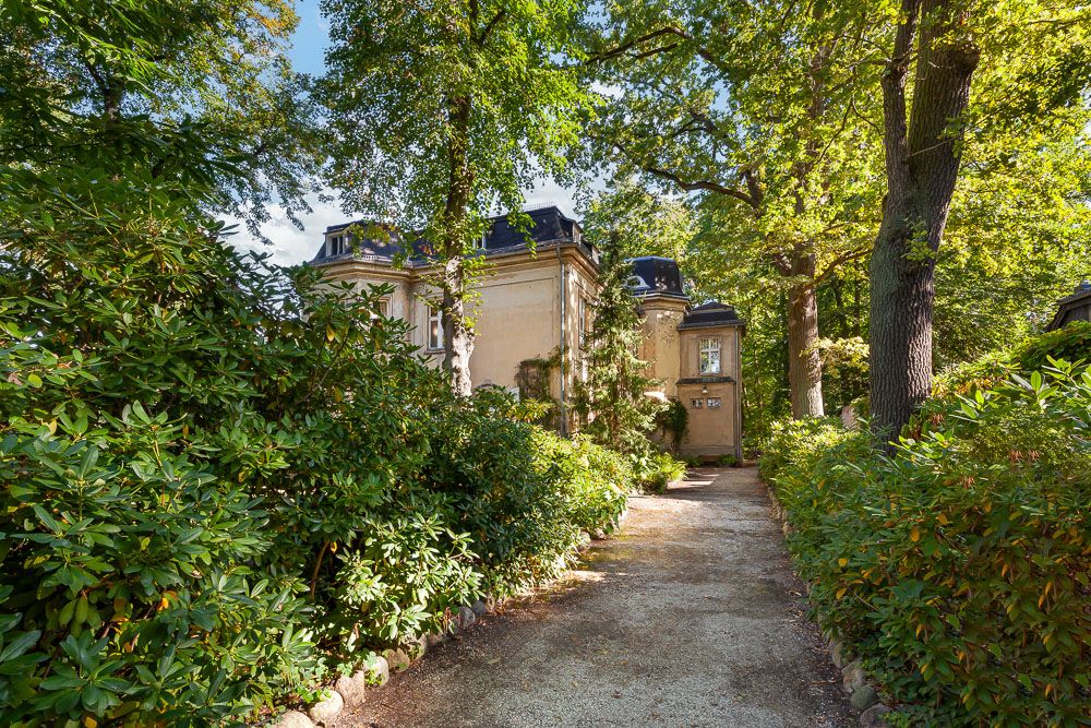 Ein schmaler Weg führt durch einen grünen Garten zu einem hellen Wohnhaus mit Bäumen im Vordergrund.
