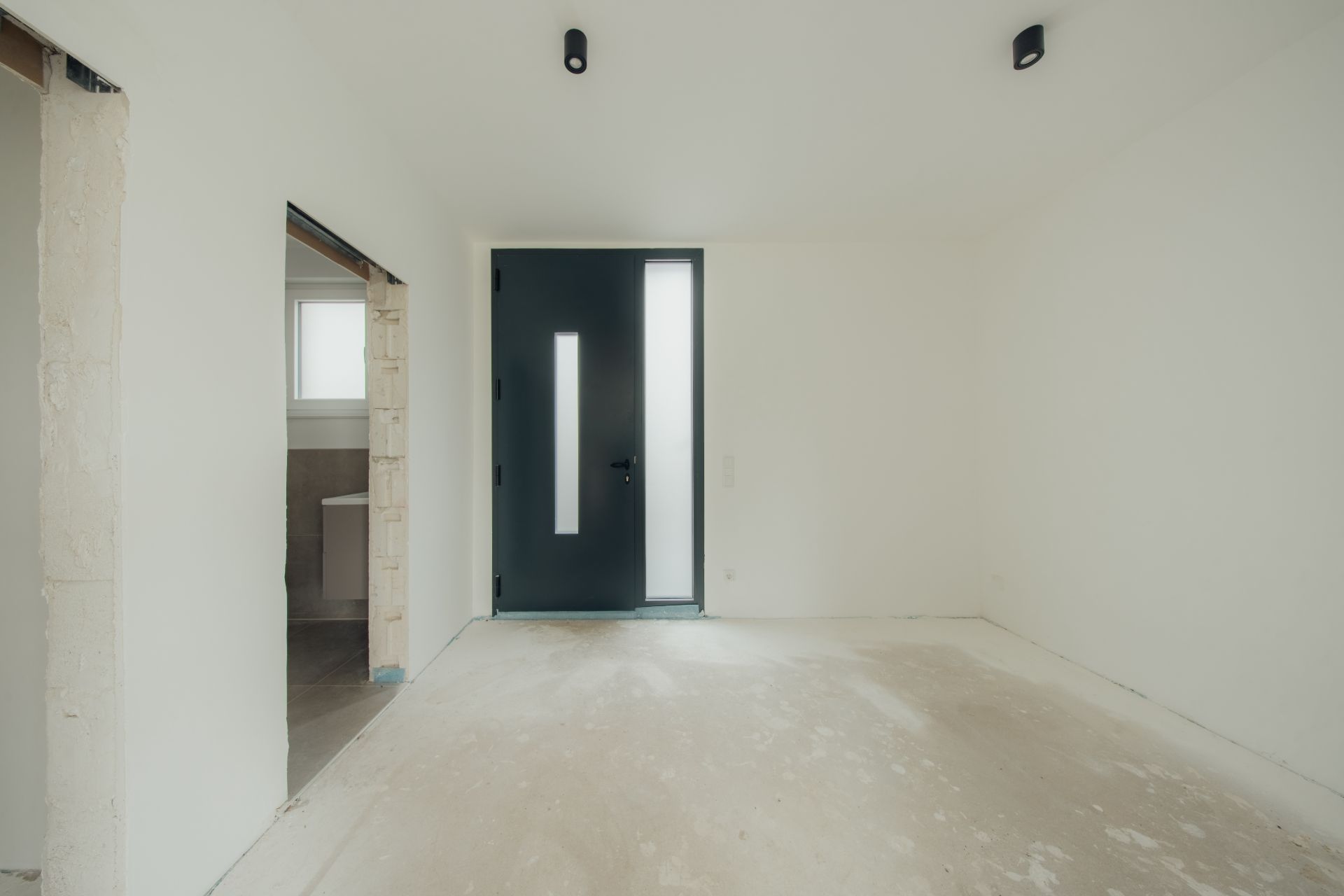 Leerer Raum mit heller Wand, dunkler Eingangstür und seitlichem Fenster, sichtbarem Rohboden.