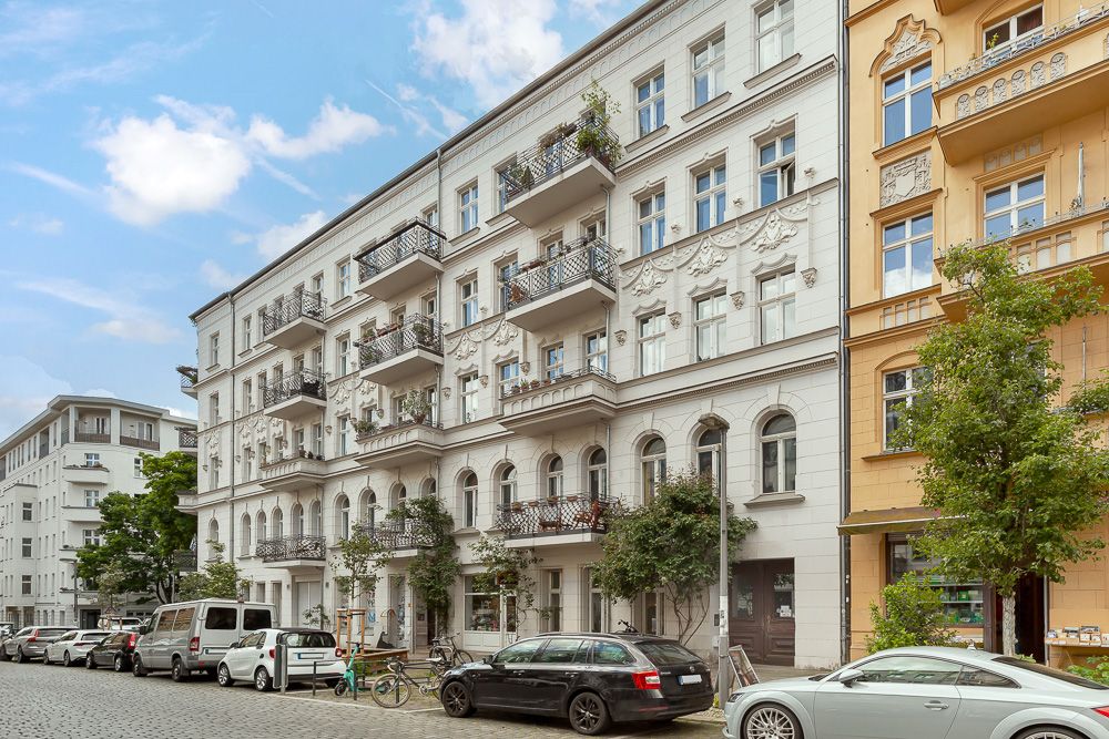 Mehrstöckiges Wohnhaus mit Balkonen an einer ruhigen Straße, Autos und Fahrräder davor geparkt.