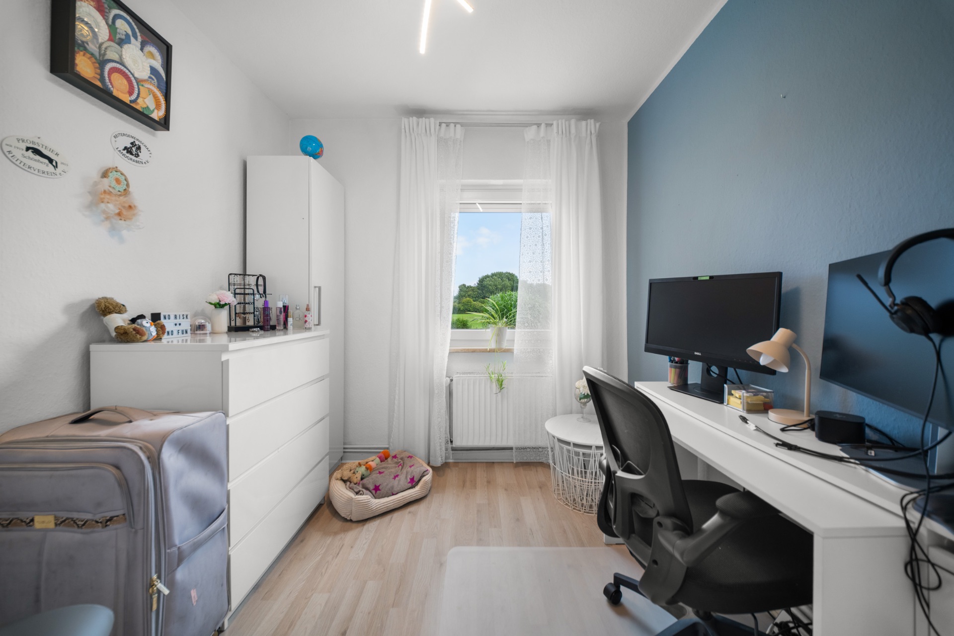 Modernes Arbeitszimmer mit Schreibtisch, Monitor, Stuhl, Kommode, Fenster und Hundebett.