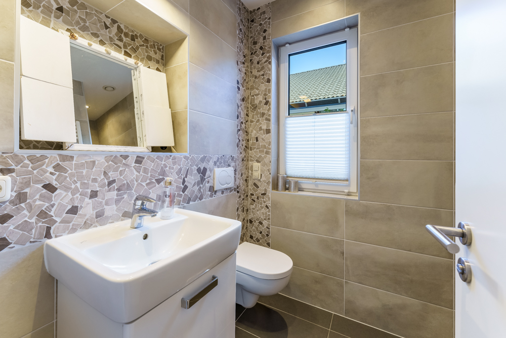 Modernes Badezimmer mit Waschbecken, Spiegel, WC und Fenster, beige Fliesen und Mosaikdekor.