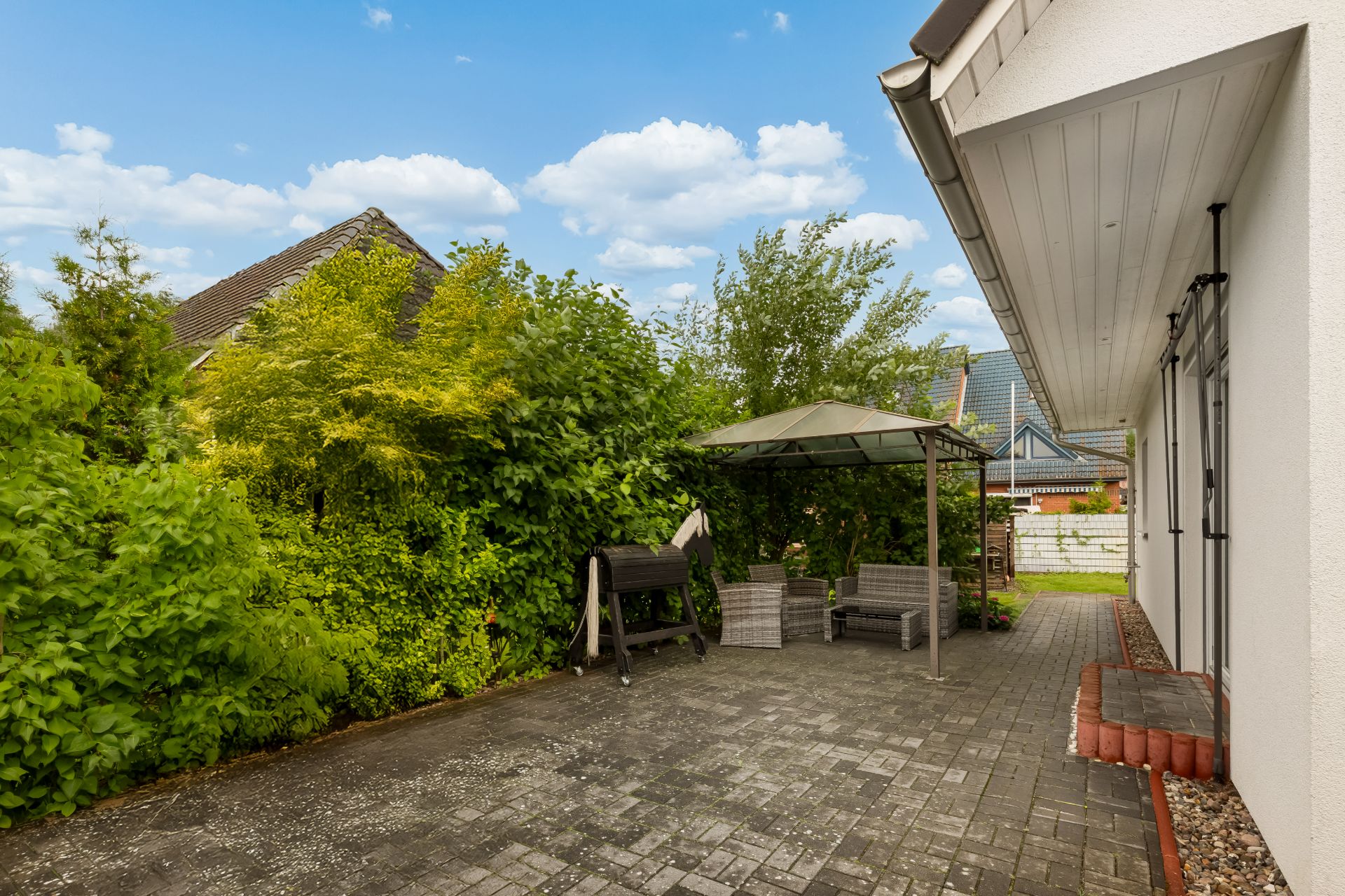 Gepflasterte Terrasse mit Sitzgruppe und Grill unter Pavillon, umgeben von viel Grün.