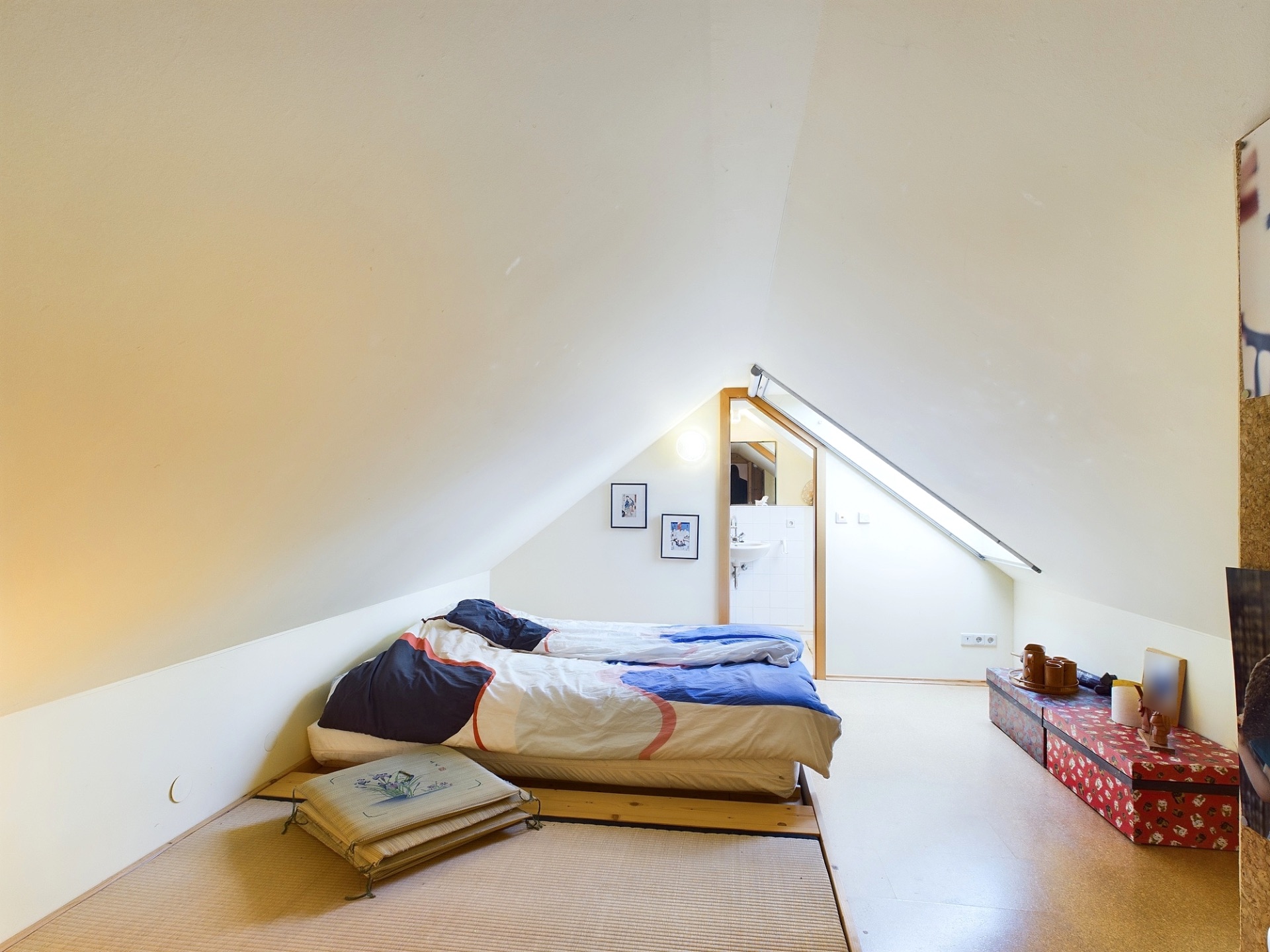 Zimmer mit schräger Decke, zwei Betten, Kissen und Kisten auf dem Boden.