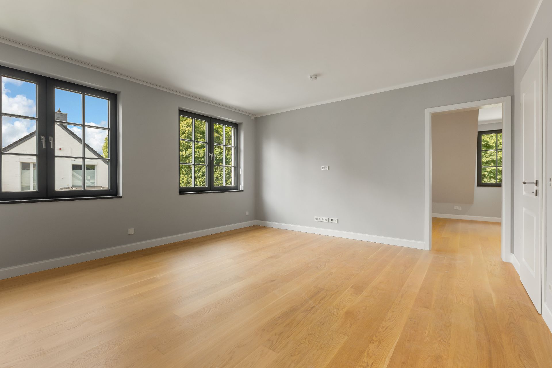 Helles, leeres Zimmer mit Holzboden und zwei großen Fenstern, angrenzend an ein weiteres Zimmer.