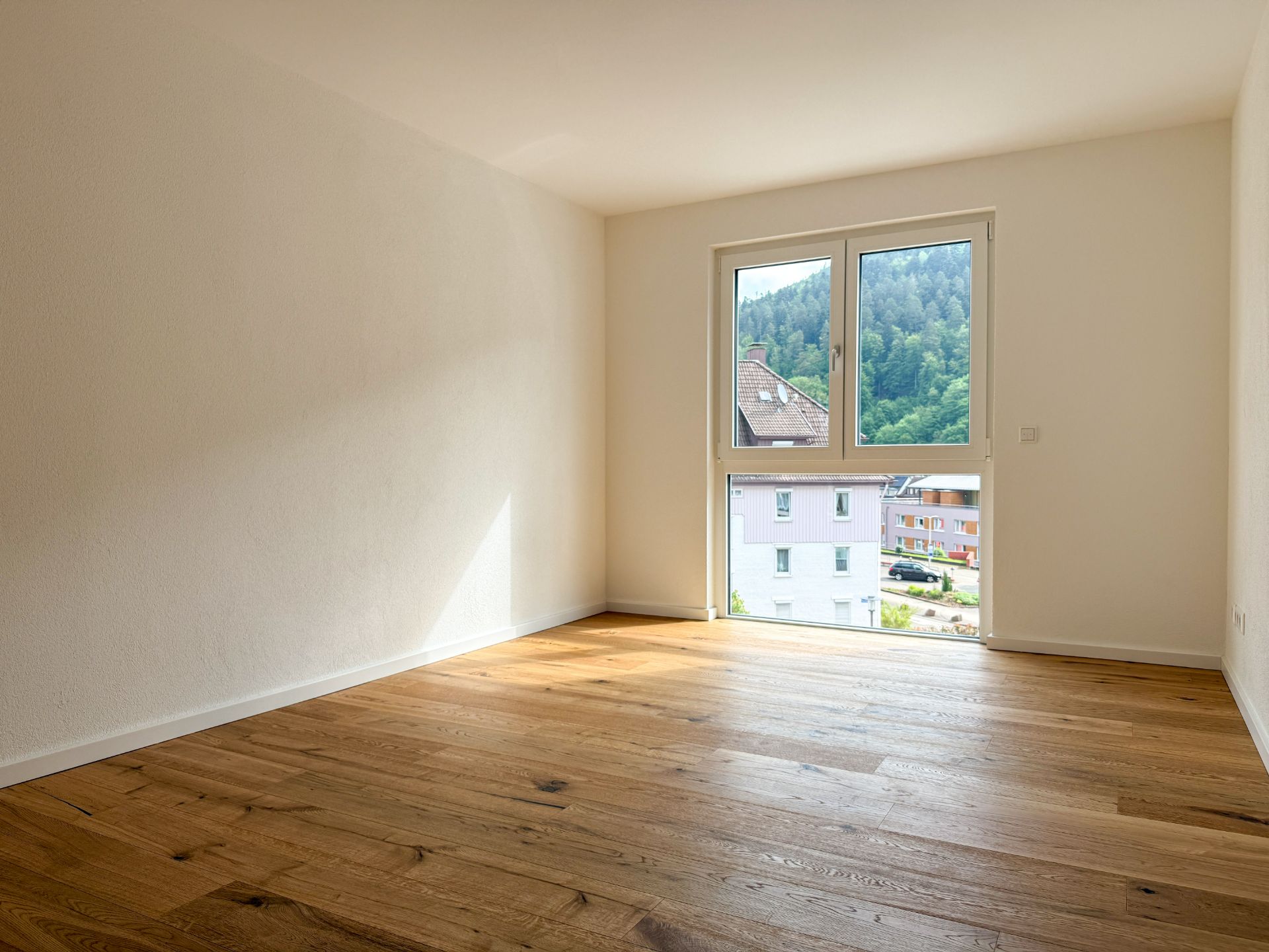 Heller, leerer Raum mit Holzfußboden und großem Fenster mit Blick auf Häuser und Bäume.