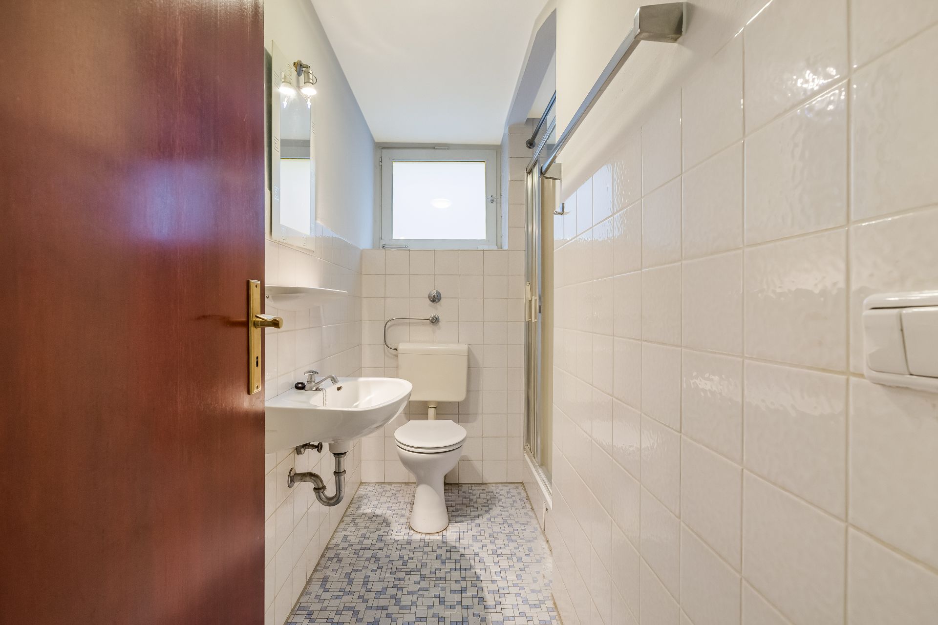 Kleines Bad mit WC, Waschbecken, Dusche und Fenster, gefliestem Boden und Wänden.