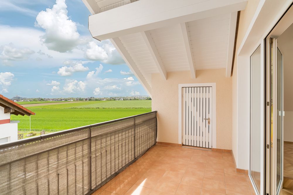 Überdachter Balkon mit Fliesenboden und Tür, Blick auf grüne Felder und Himmel.