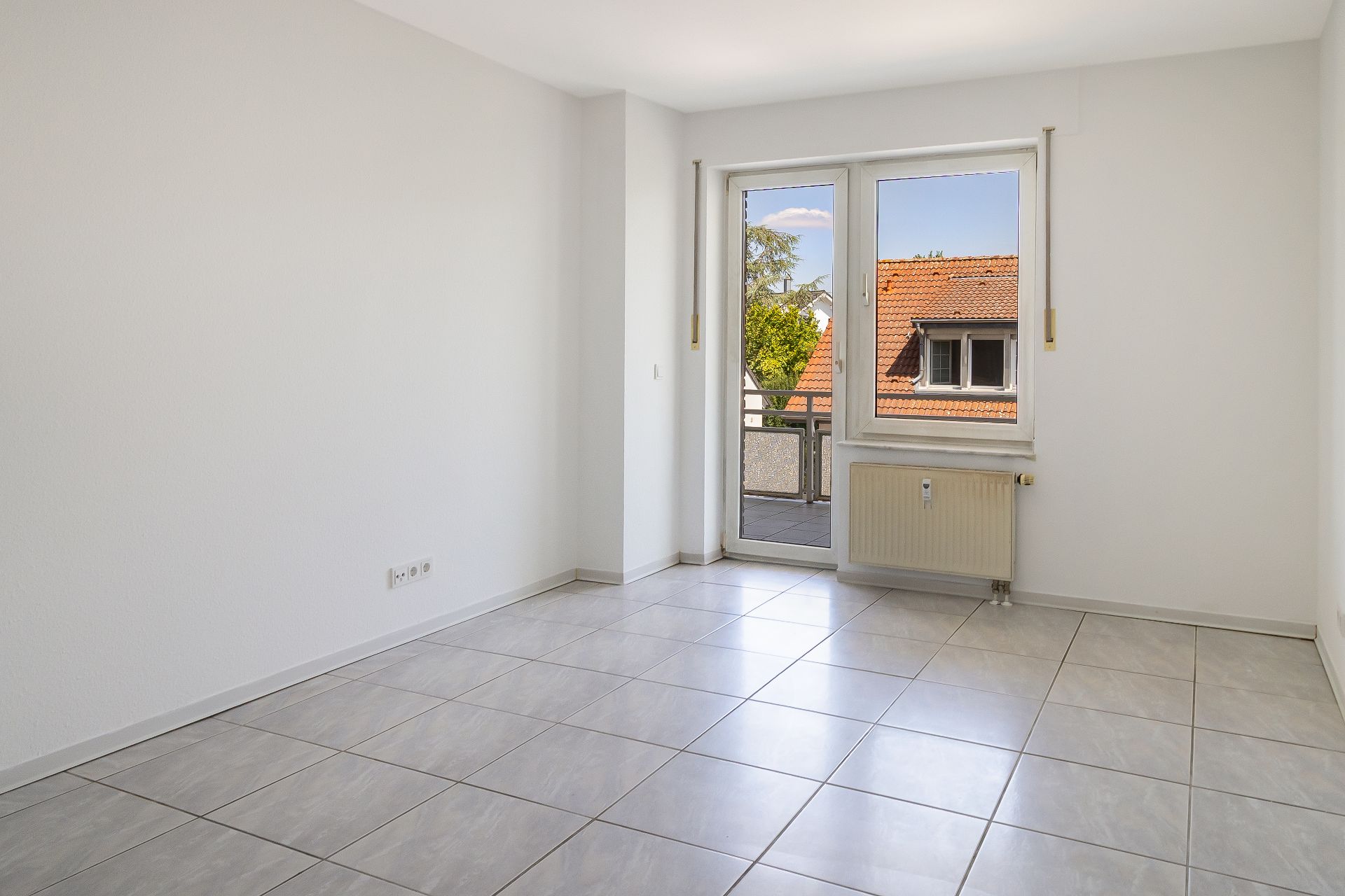 Leerer, heller Raum mit Fliesenboden und einem Fenster mit Blick auf Häuserdächer.