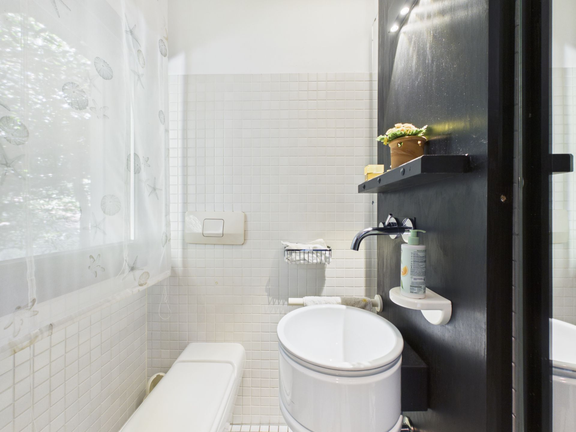 Kleines Badezimmer mit Fenster, WC, Waschbecken und schwarzen Wandregalen.