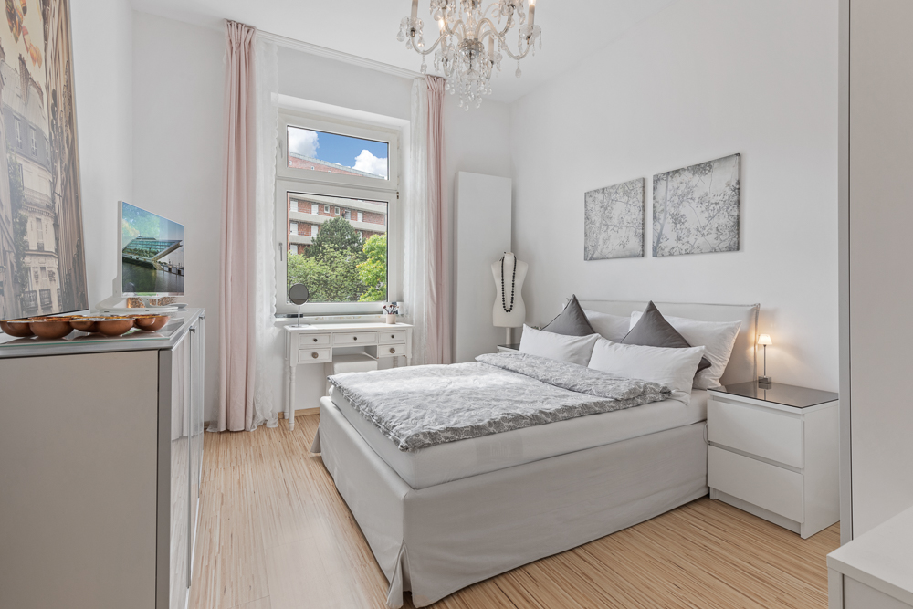 Helles Schlafzimmer mit Doppelbett, Schreibtisch am Fenster und Kronleuchter.