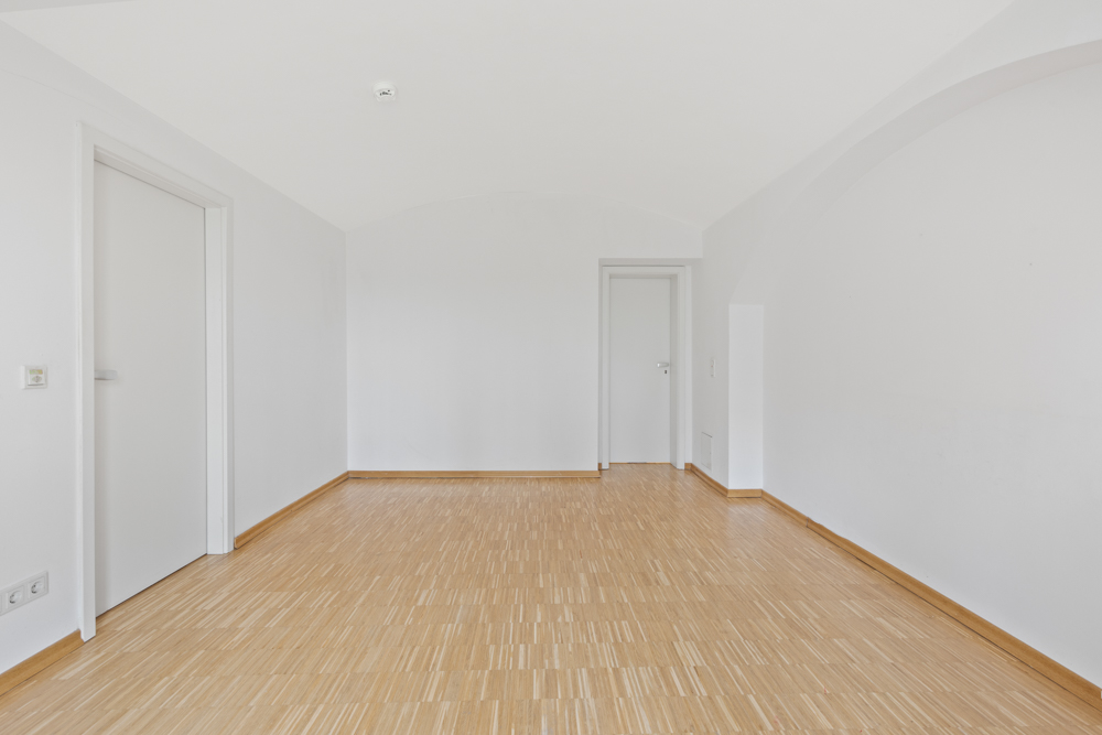 Leerer Raum mit hellem Holzboden und weißen Wänden, zwei geschlossene Türen.