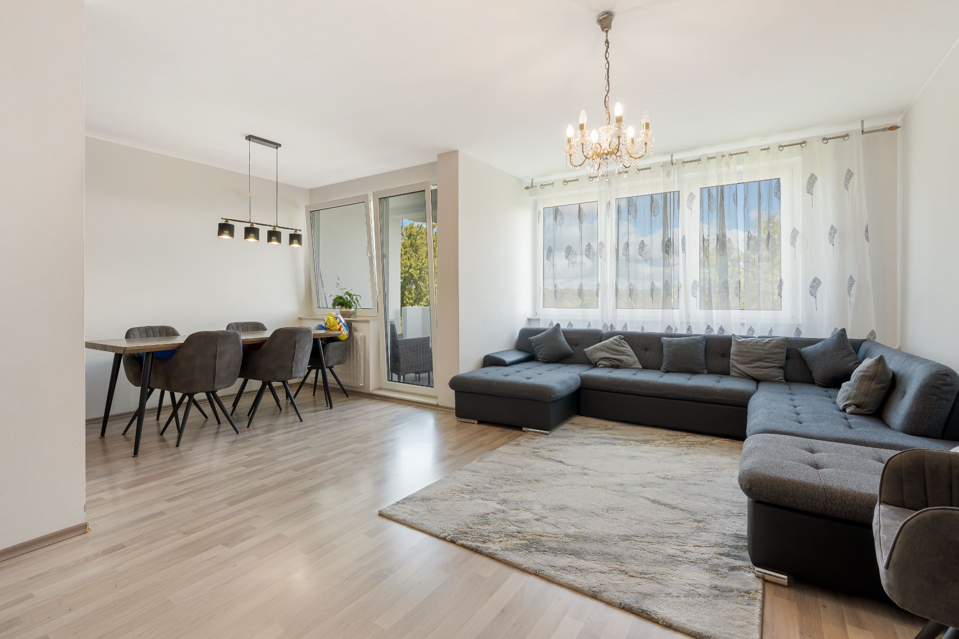 Helles Wohnzimmer mit großer grauer Eckcouch, Esstisch und großen Fenstern mit Vorhängen.