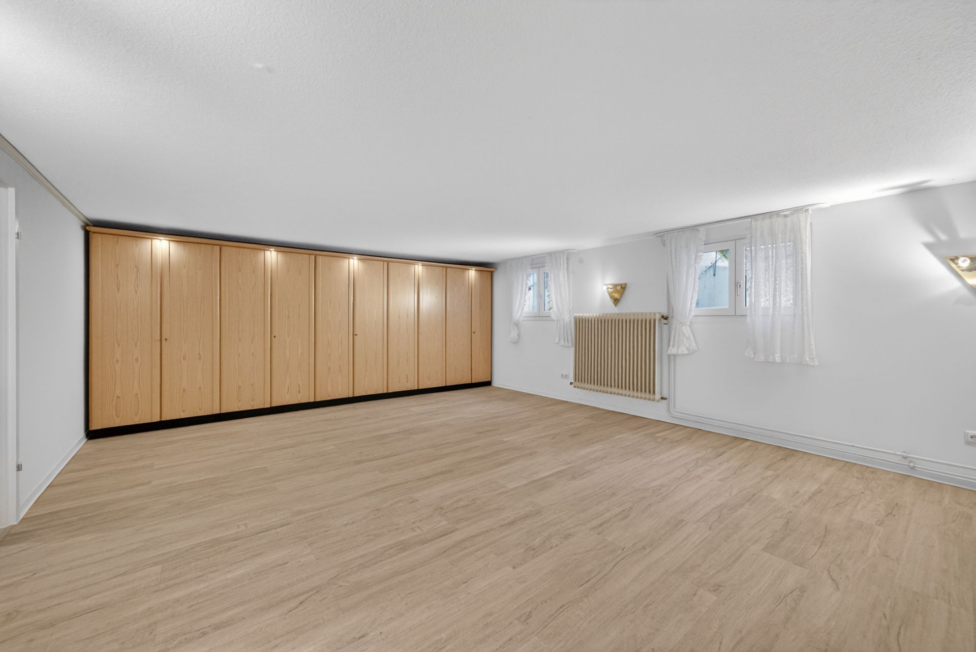 Heller Raum mit Holzfußboden, großen Einbauschränken und zwei Fenstern mit weißen Vorhängen.
