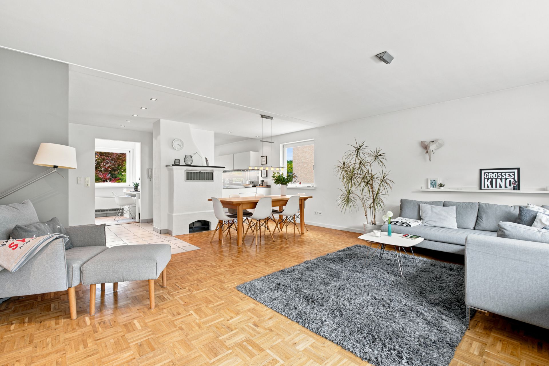 Helles Wohnzimmer mit grauem Sofa, Teppich, Essbereich und offener Küche, Parkettboden und Dekopflanze.