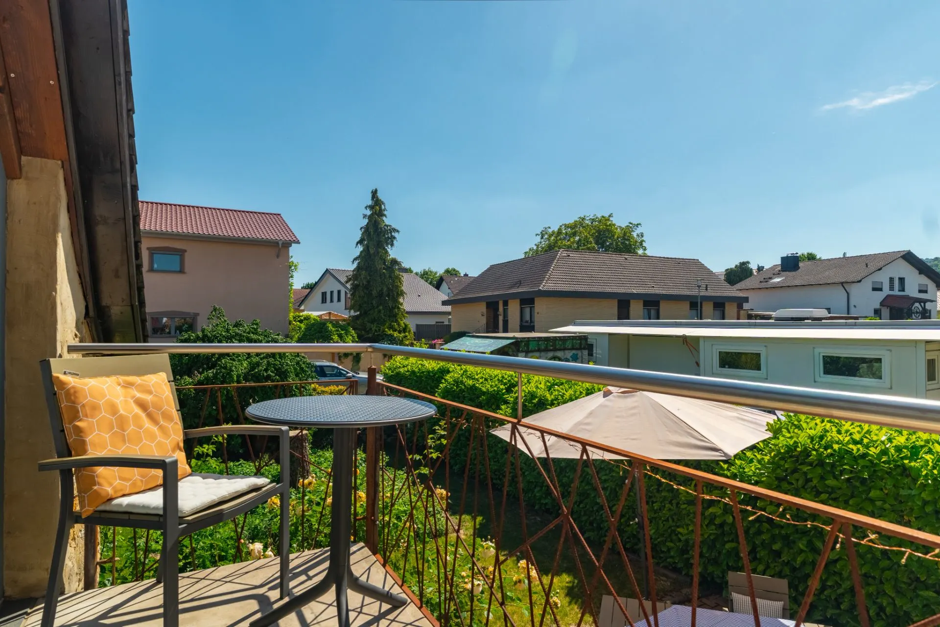 Balkon mit Stuhl, rundem Tisch und Blick auf Nachbarhäuser und Garten bei Sonnenschein.