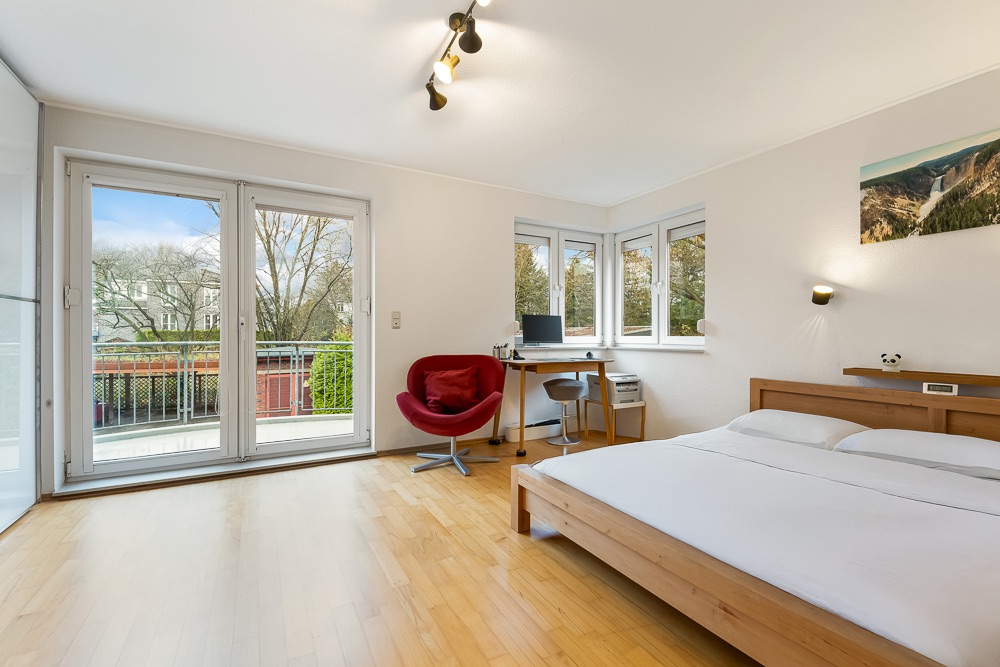 Helles Schlafzimmer mit großem Bett, Schreibtisch, rotem Sessel und Zugang zu einem Balkon.