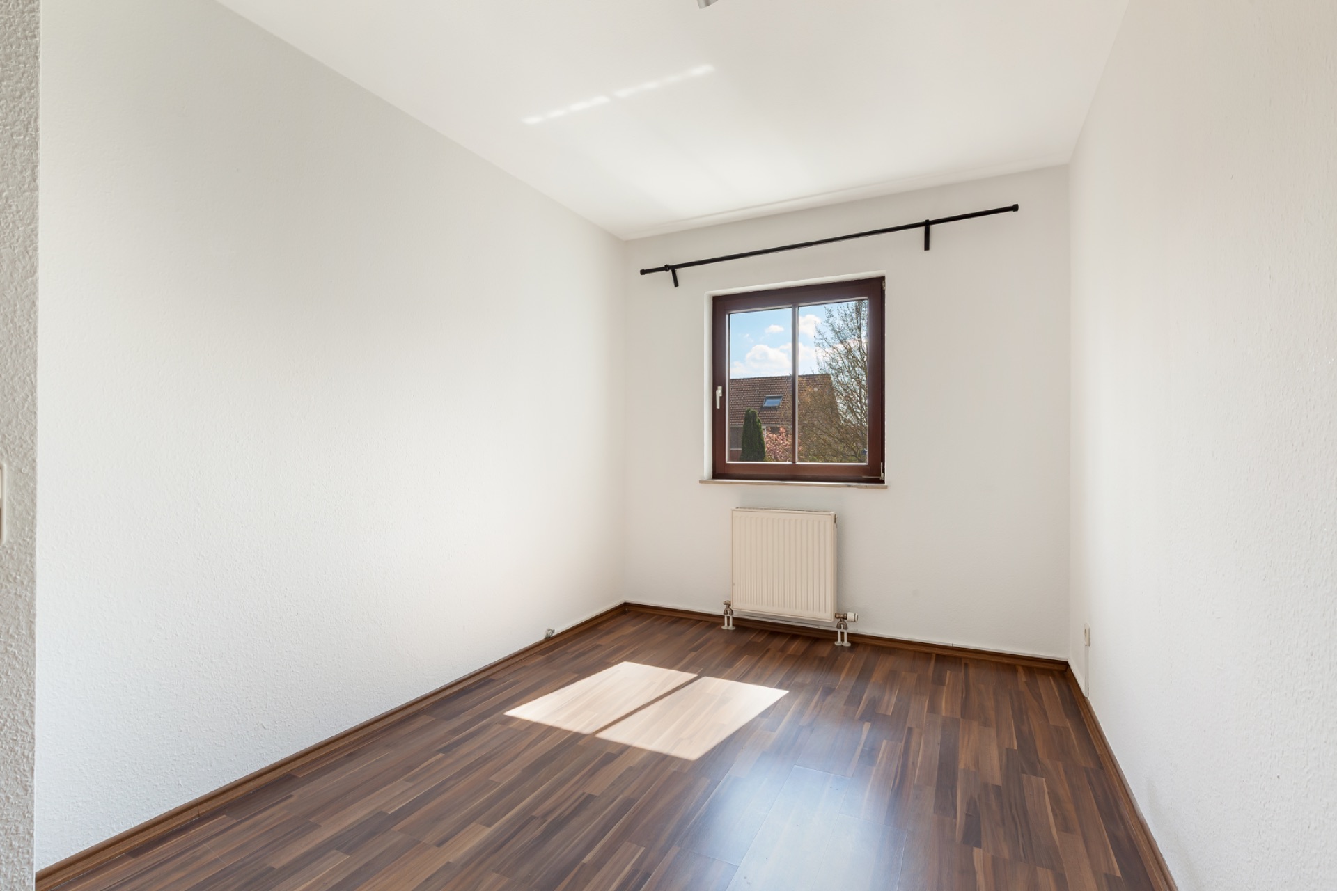 Leeres Zimmer mit braunem Holzboden, weiß gestrichenen Wänden und einem Fenster mit Heizkörper darunter.
