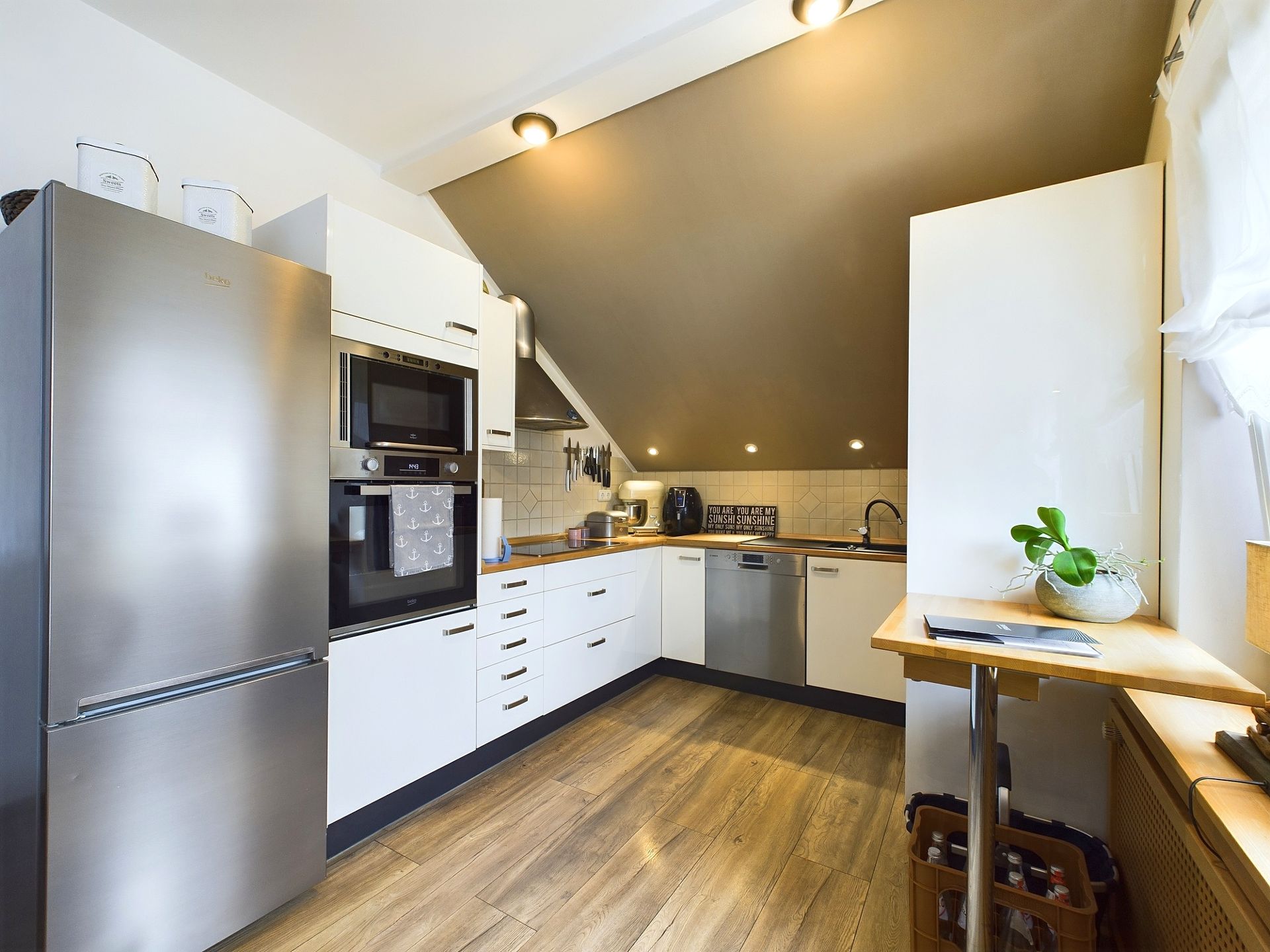 Moderne Einbauküche mit Schräge, weißen Schränken, Holzboden und Edelstahlgeräten.