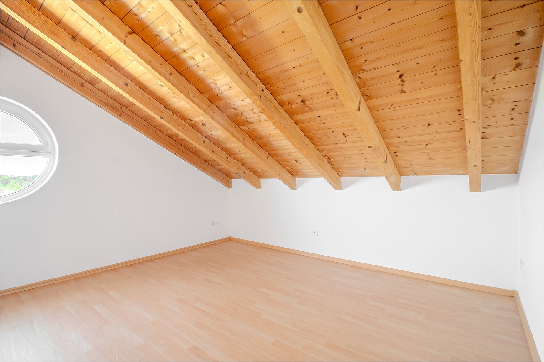 Leerer Dachraum mit schräger Holzdecke, hellem Boden und rundem Fenster an der Seitenwand.