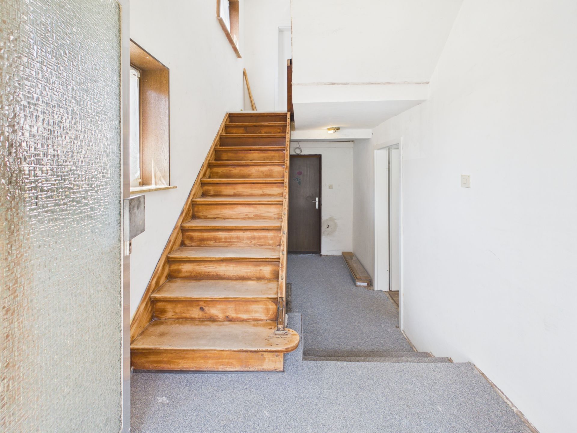 Ein Flur mit hölzerner Treppe, grauem Teppichboden und weißen Wänden.