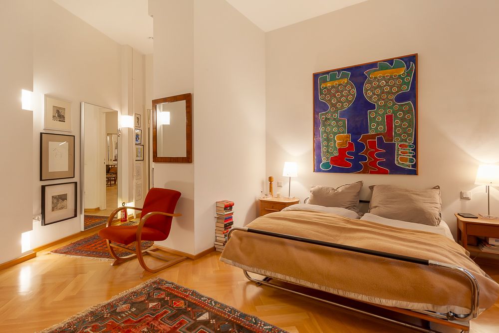 Helles Schlafzimmer mit Doppelbett, rotem Sessel, Teppich und buntem Bild an der Wand.