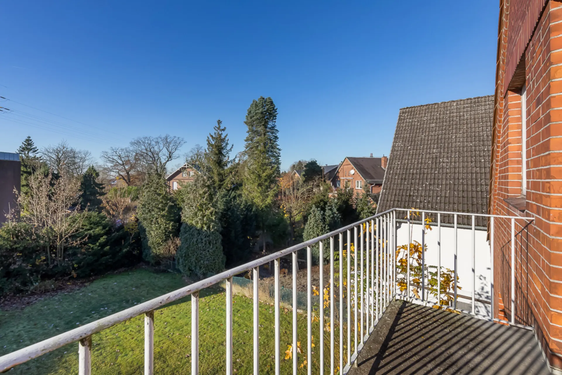 Blick von einem Balkon auf einen Garten mit Bäumen und Nachbarhäusern bei klarem Himmel.