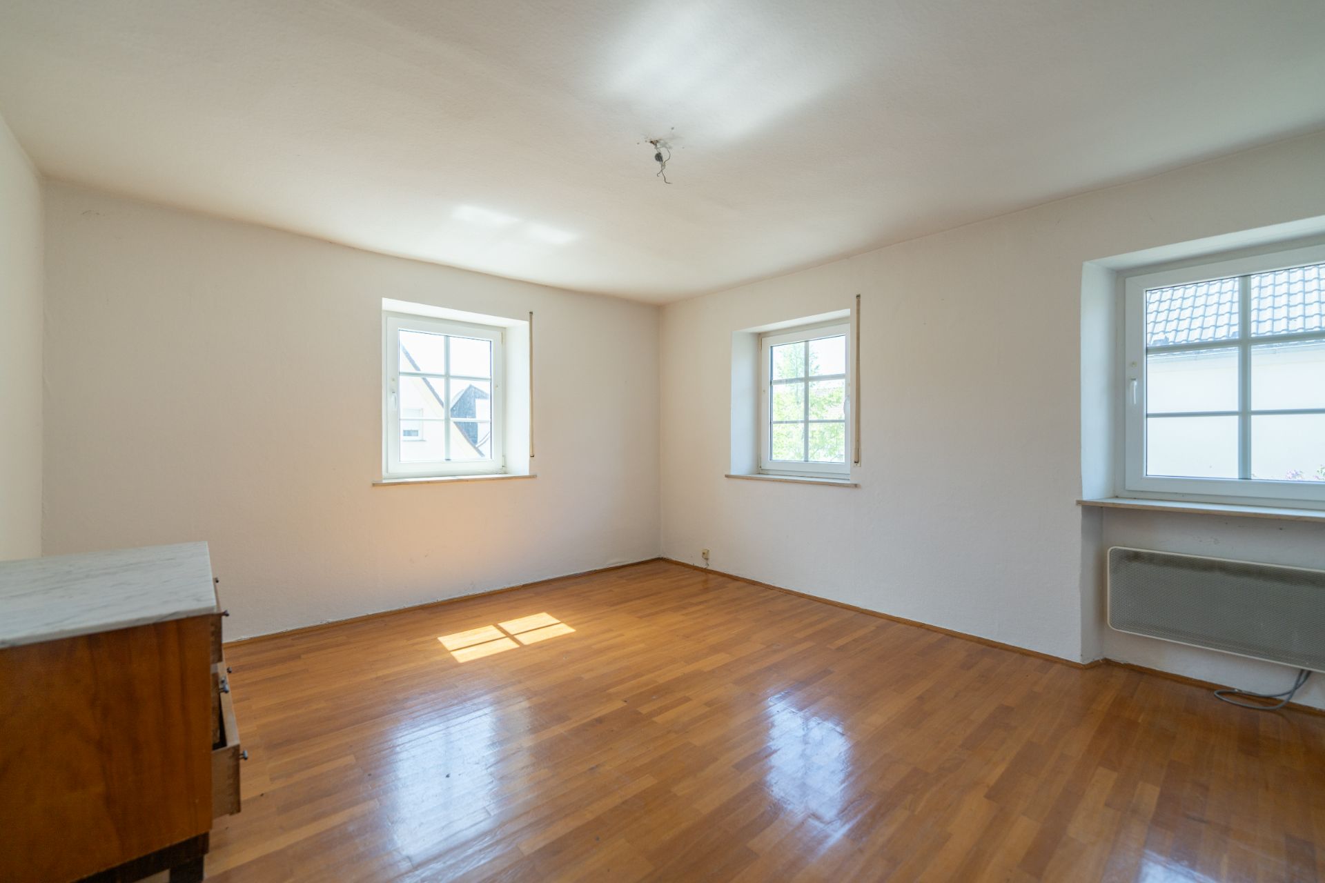 Helles Zimmer mit drei Fenstern, Parkettboden und einer Kommode an der Wand.