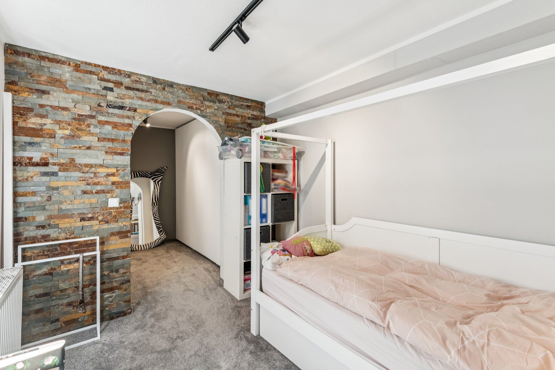 Kinderzimmer mit weißem Bett, Steinwand und offenem Regal neben einem Rundbogen.