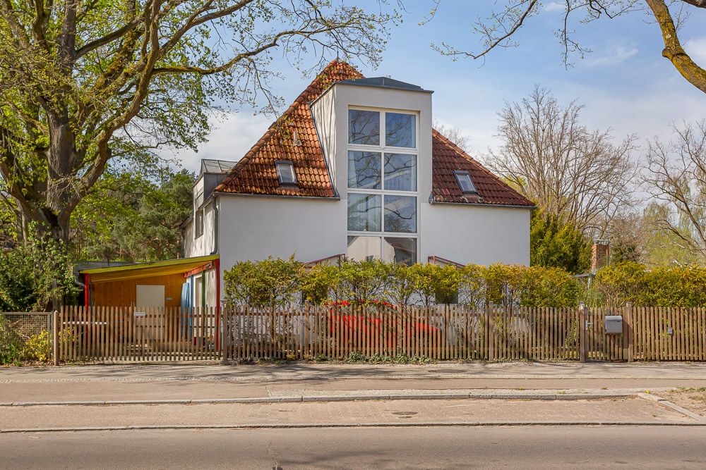 Modernes Einfamilienhaus mit rotem Satteldach und großem Fenster, umzäunt von Hecken und Holzzaun.