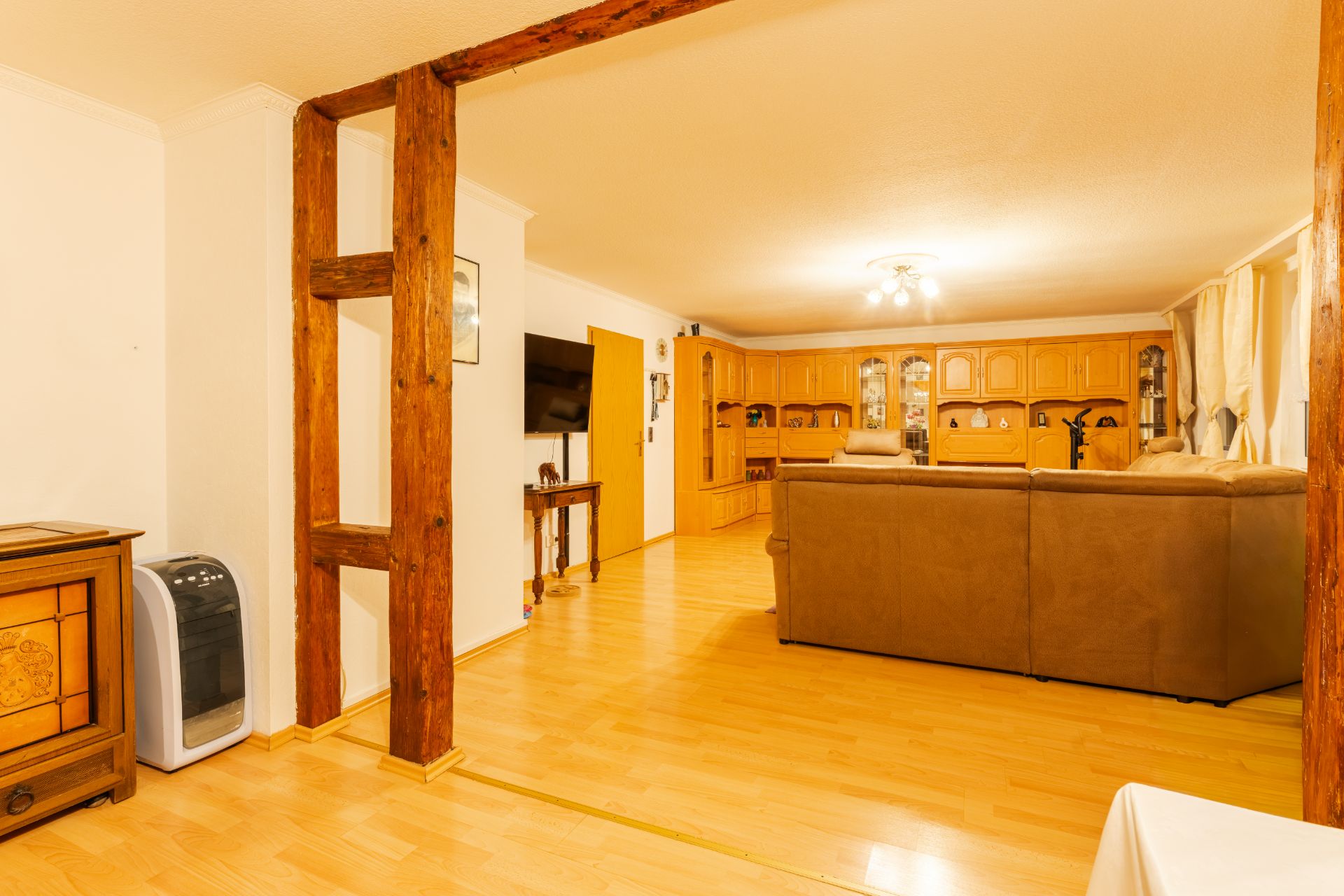 Wohnzimmer mit Holzbalken, branem Sofa, Holzschrankwand und Fernseher auf hellem Holzboden.