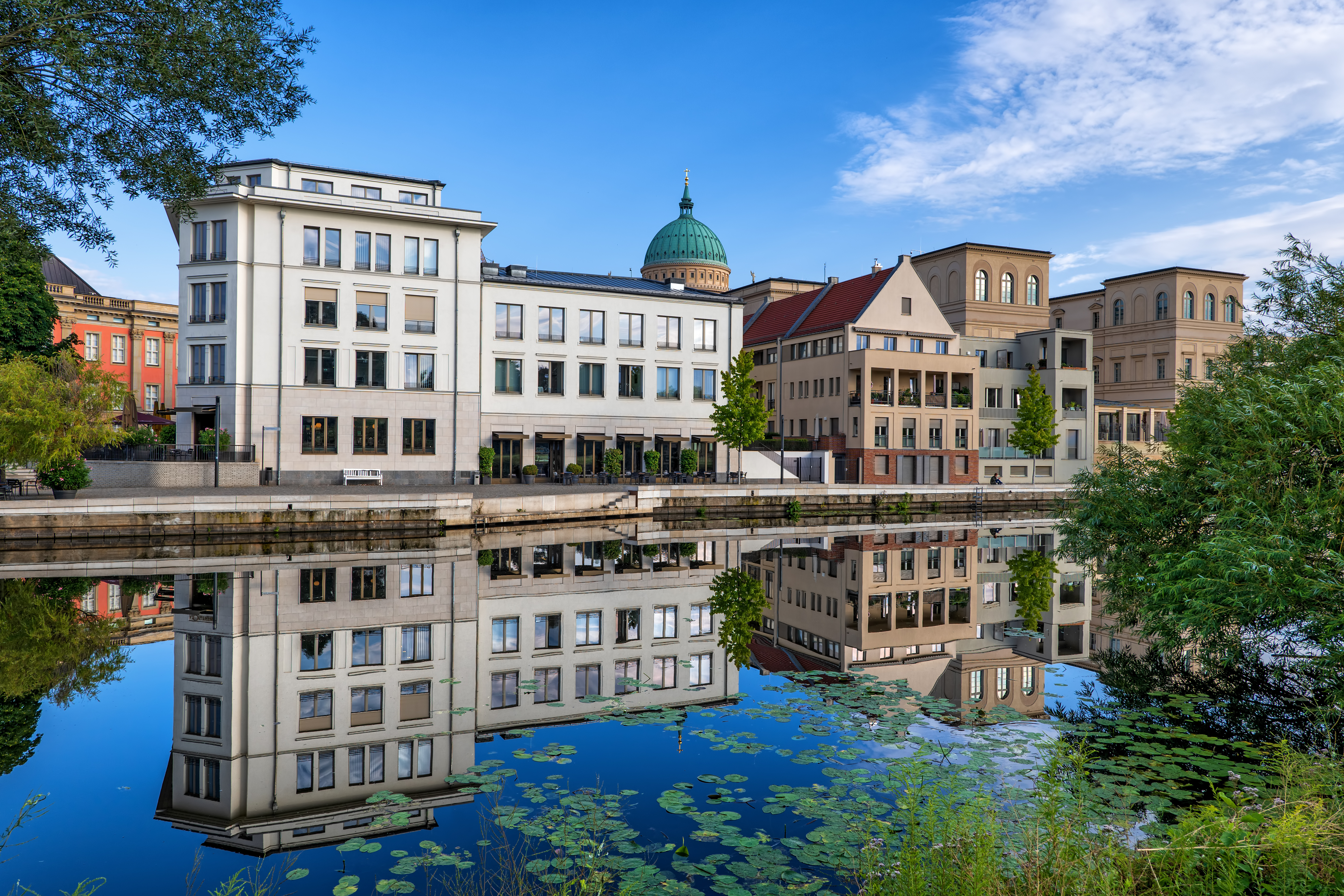 Mehrstöckige Wohnhäuser spiegeln sich in einem ruhigen Fluss, vorne Seerosen, blauer Himmel.