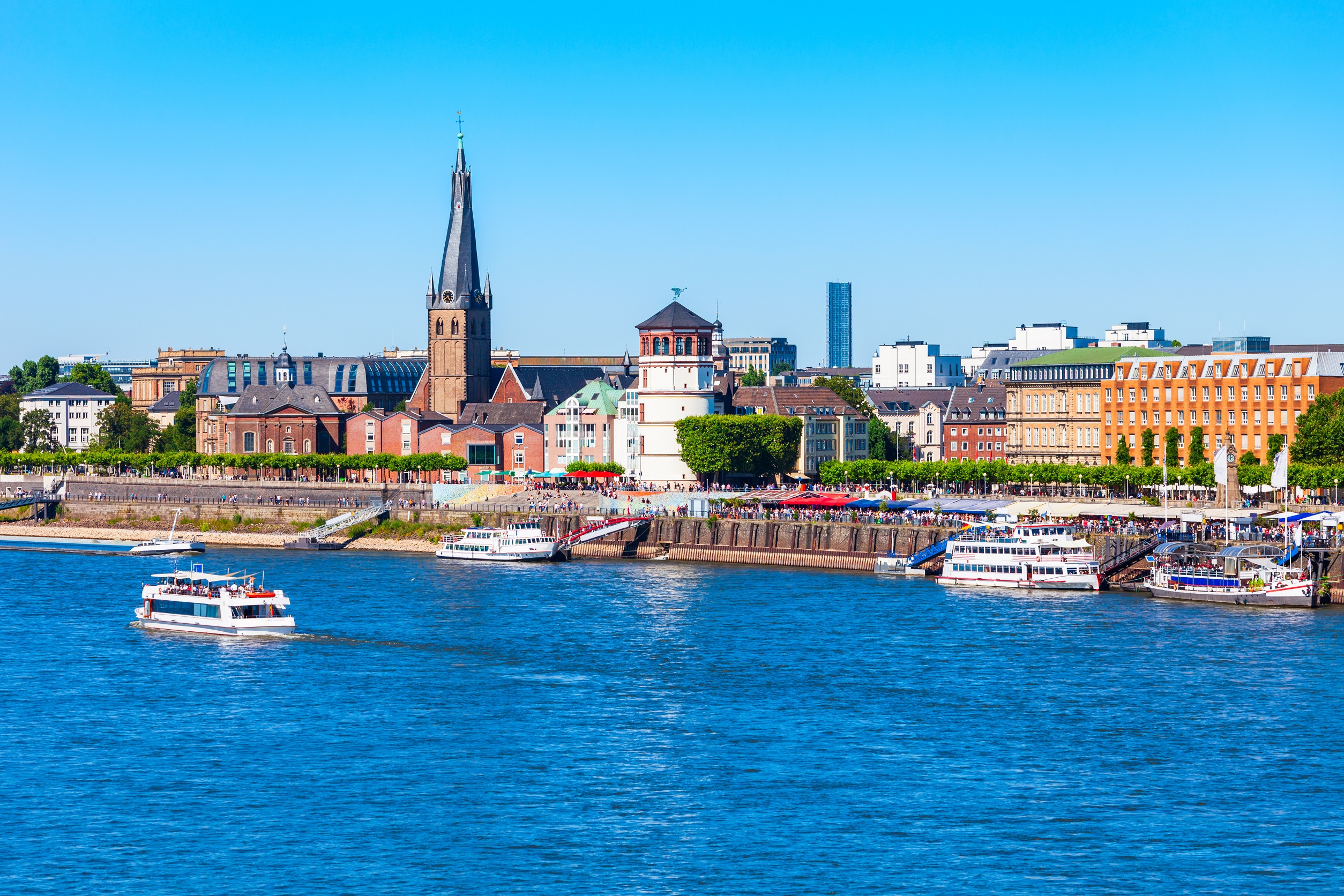 Stadtpanorama mit Kirche, historischen Gebäuden und Schiffen am Flussufer bei sonnigem Wetter.