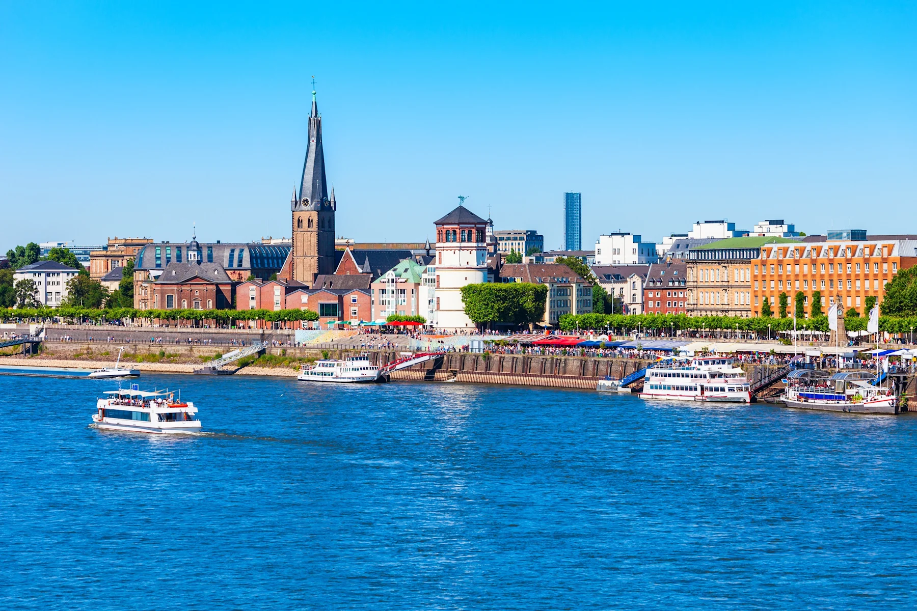 Stadtpanorama mit Kirche, historischen Gebäuden und Schiffen am Flussufer bei sonnigem Wetter.