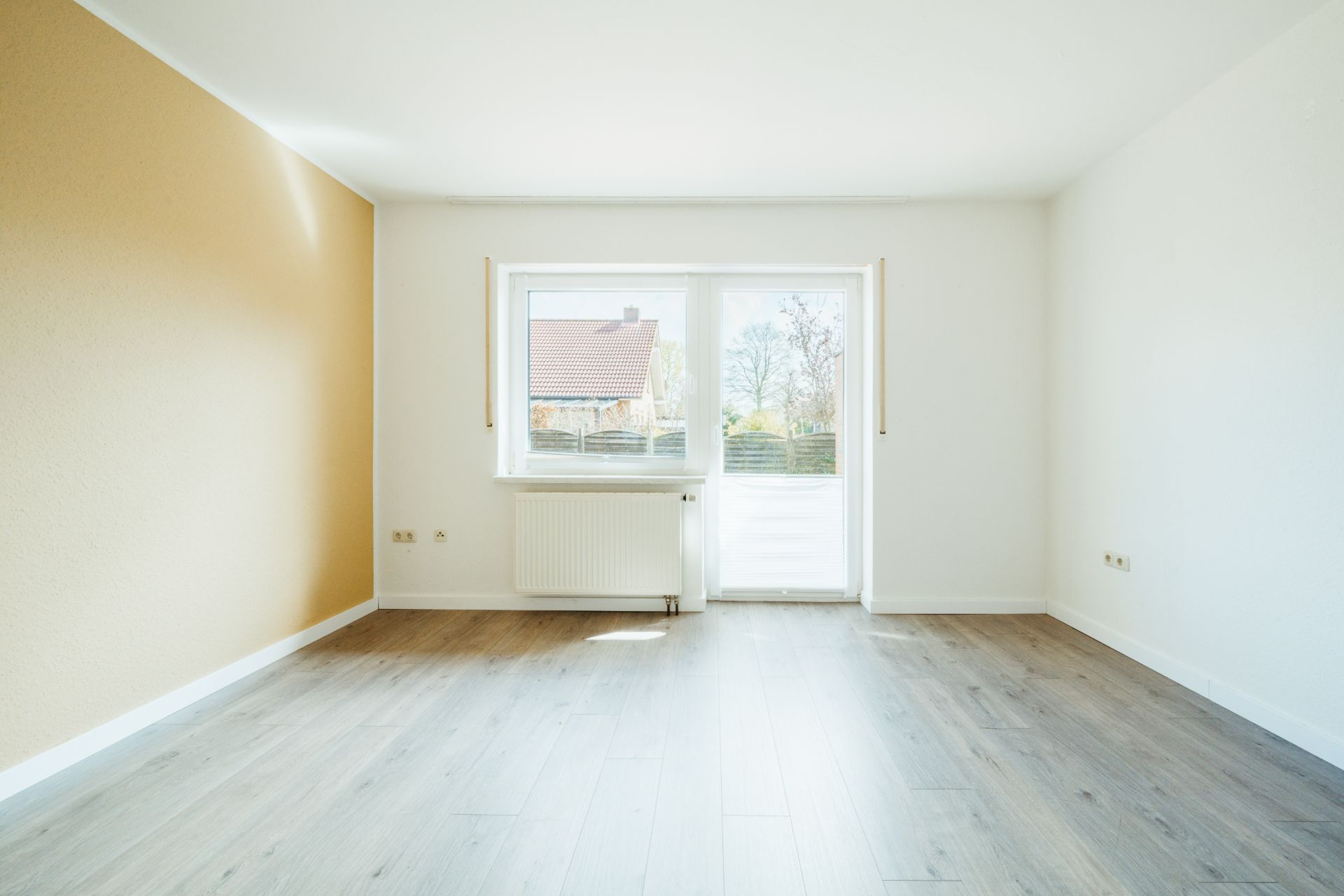 Heller leerer Raum mit Holzfußboden, großem Fenster und Tür zu einer Terrasse.