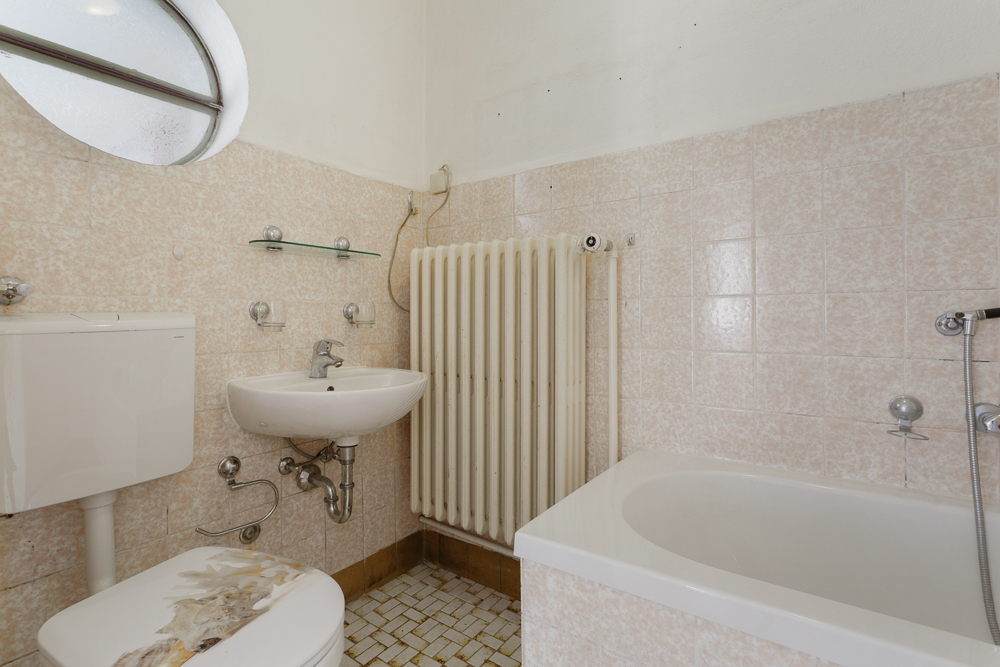Kleines Bad mit Toilette, Waschbecken, Badewanne, Heizkörper und rundem Fenster.