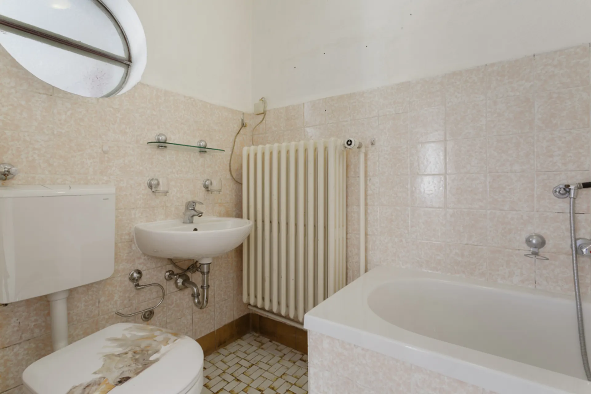 Kleines Bad mit Toilette, Waschbecken, Badewanne, Heizkörper und rundem Fenster.