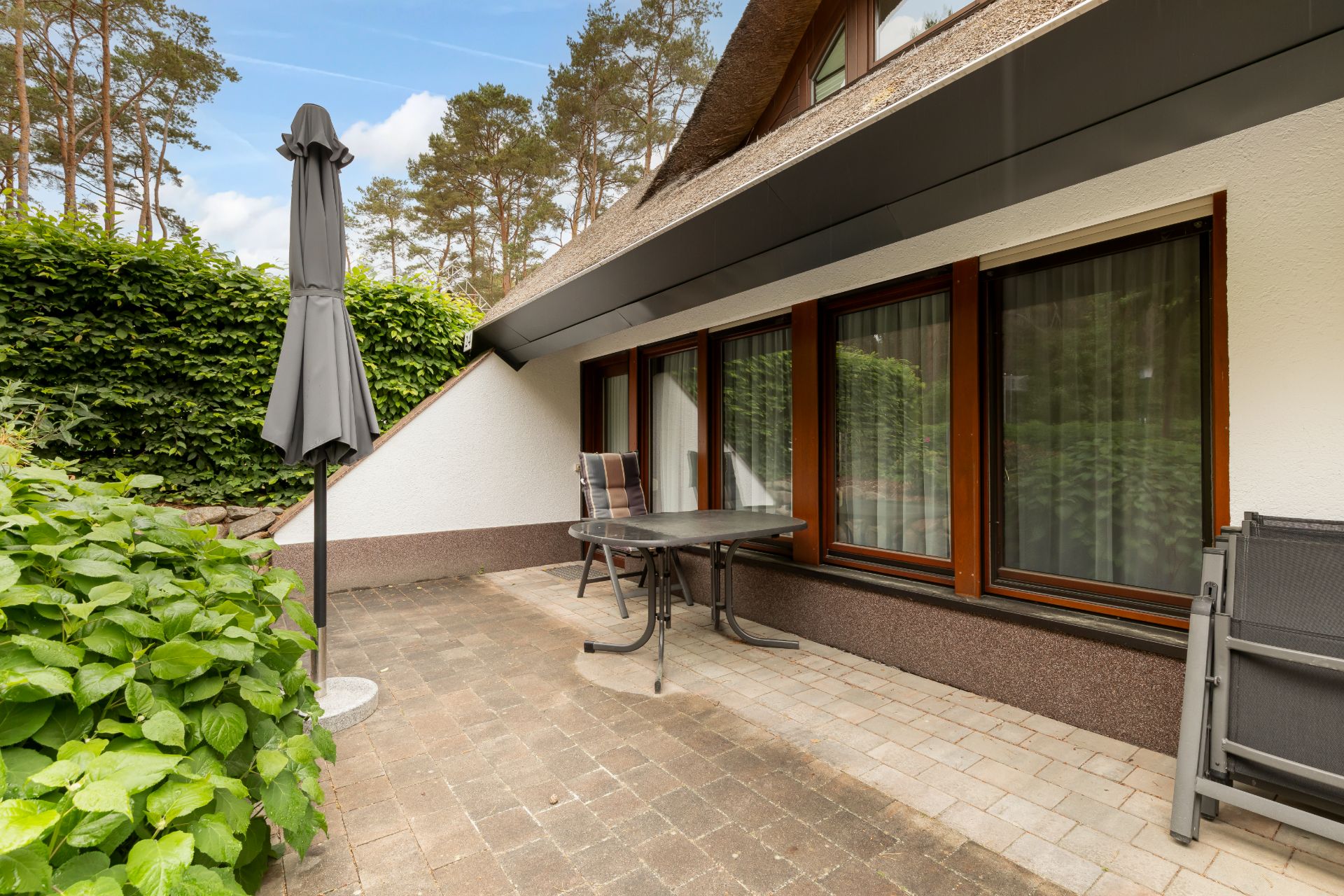 Terrasse mit Tisch, Stühlen und geschlossener Sonnenschirm, neben Haus mit großen Fenstern.