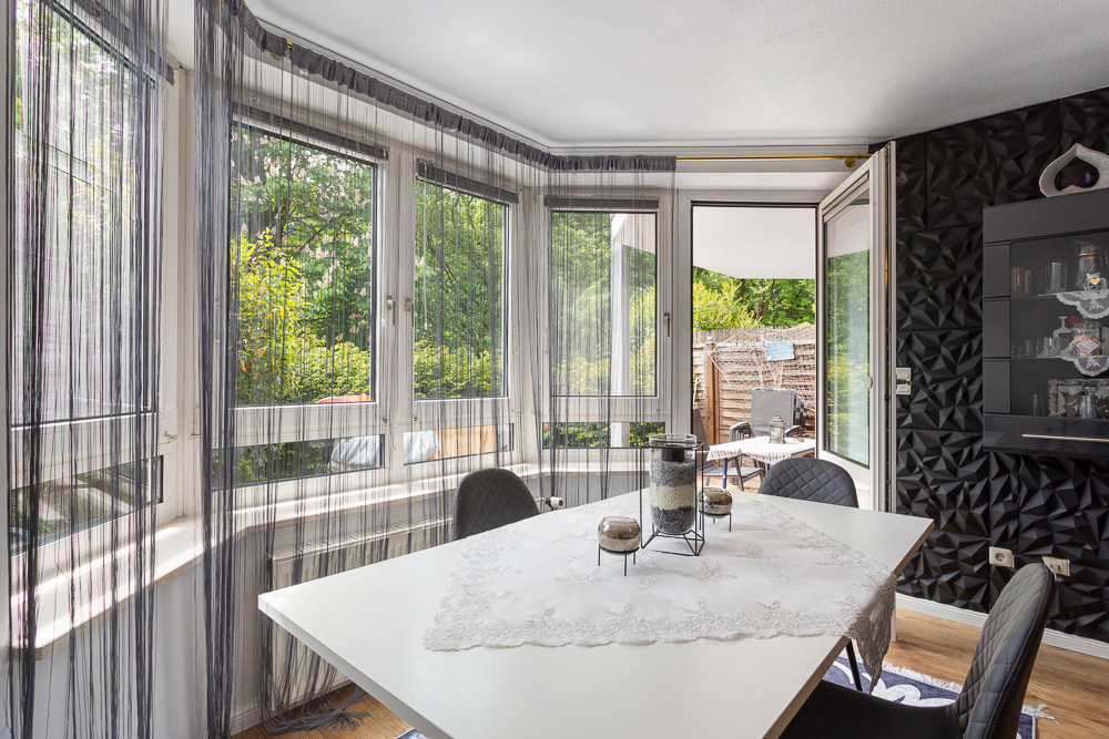 Essbereich mit weißem Tisch, vier Stühlen, großen Fenstern und Blick auf den Garten.