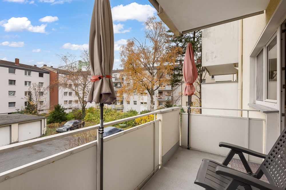 Balkon mit zwei geschlossenen Sonnenschirmen und einem Stuhl, Blick auf Wohnhäuser und Bäume.
