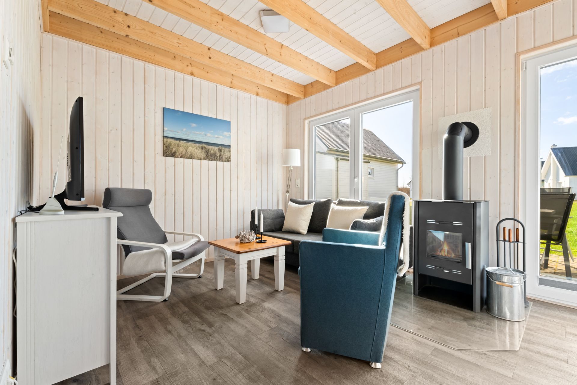 Wohnzimmer mit Holzbalkendecke, Sofa, Sessel, Fernseher und Kamin, große Fenster mit Blick nach draußen.