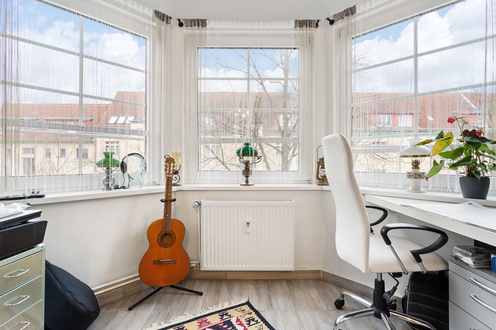 Helles Arbeitszimmer mit Schreibtisch, weißem Stuhl, Gitarre und großen Fenstern.