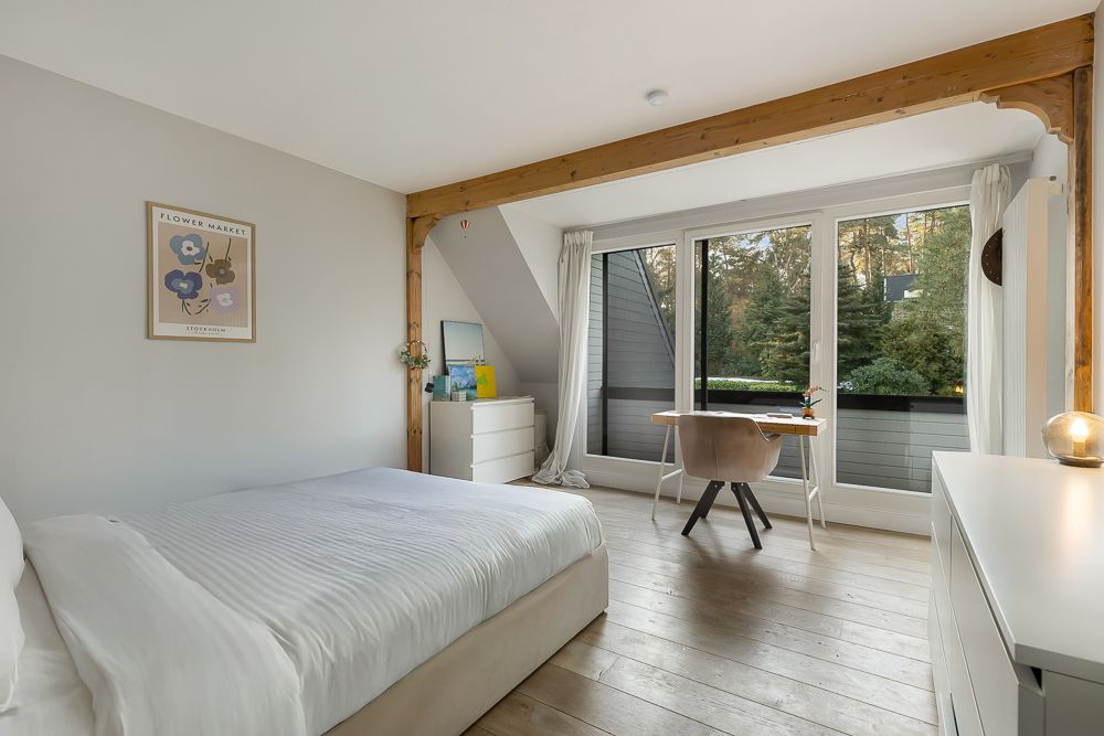 Helles Schlafzimmer mit Bett, Schreibtisch, Kommode und großem Fenster mit Blick ins Grüne.