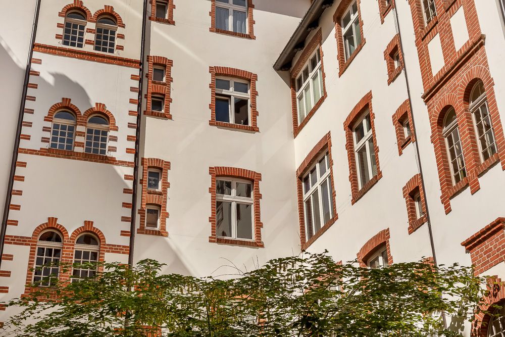 Innenhof eines Wohnhauses mit roten Backsteinfenstern und weißen Wänden, unten Bäume sichtbar.