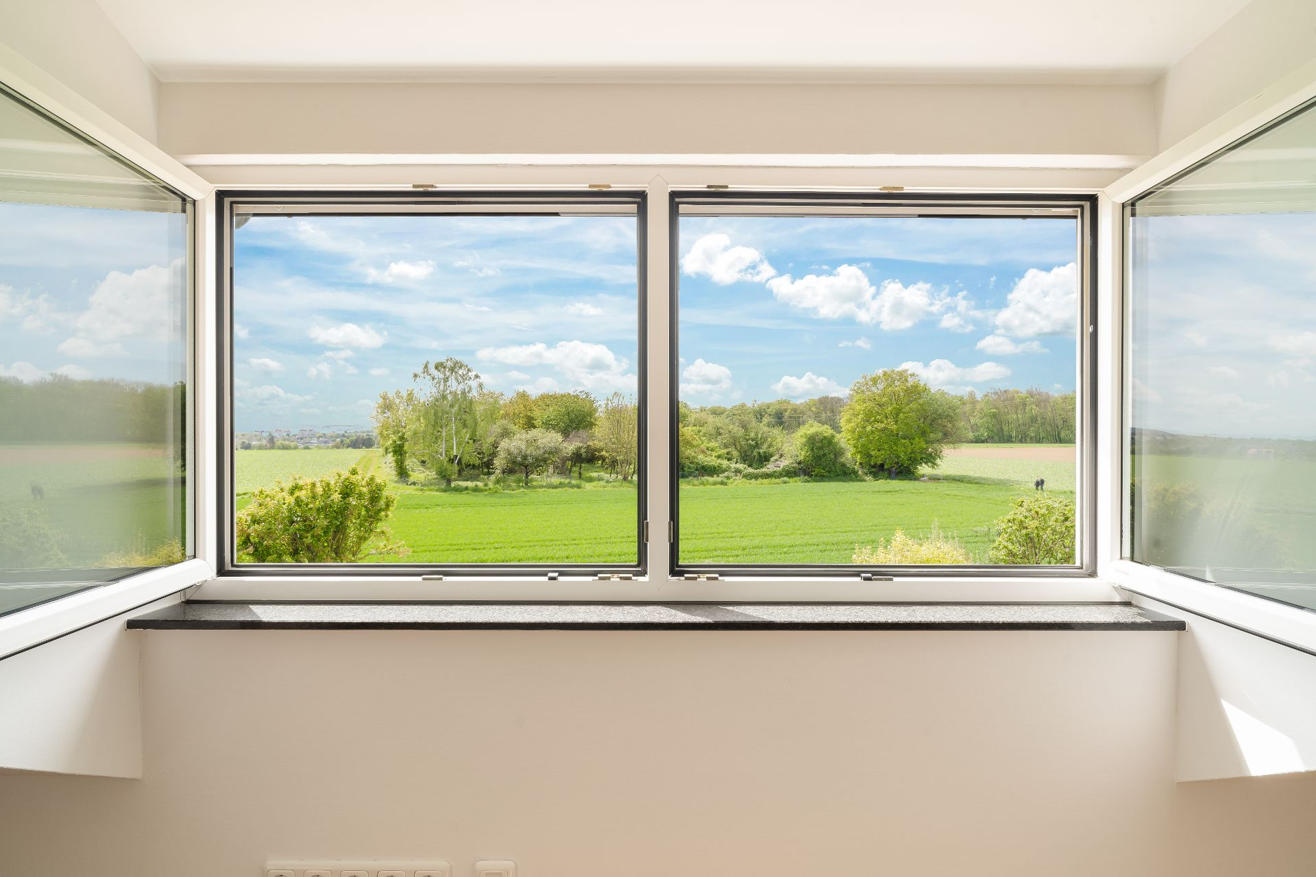 Großes offenes Fenster mit Blick auf grüne Felder und Bäume unter blauem Himmel.