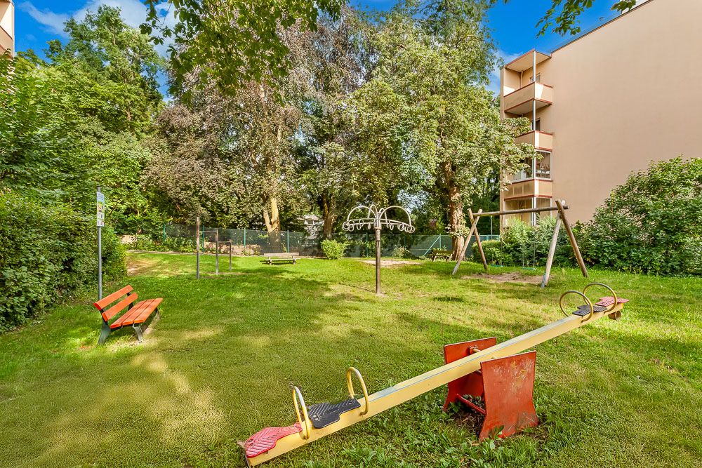 Grüner Spielplatz mit Wippe, Schaukel, Bank und umliegenden Bäumen neben Wohngebäude.