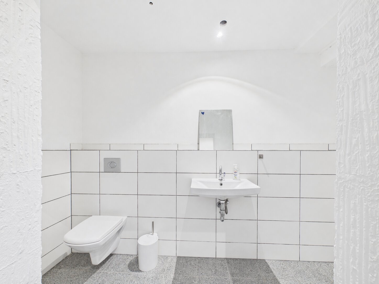 Modernes, weiß gefliestes Badezimmer mit Wand-WC, Waschbecken und kleinem Spiegel.